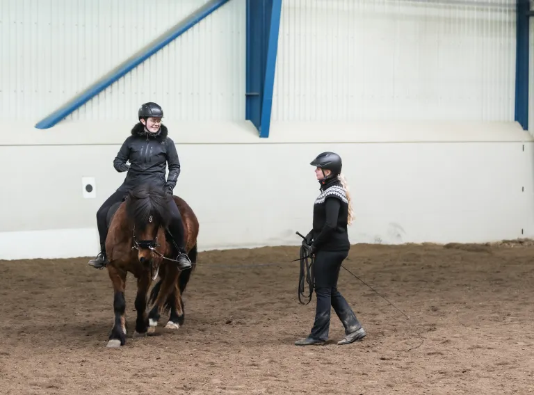 A rider wearing a helmet rides an Icelandic horse in an indoor riding arena while another helmeted person stands nearby providing instruction with a training line.