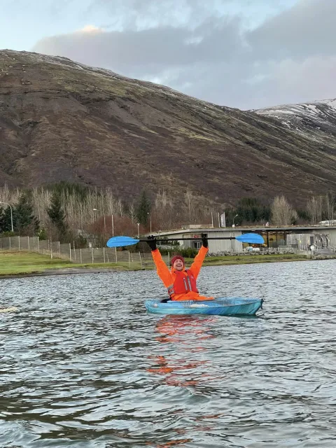 Viktor orri out on water in a kayak, holding his paddle above head like he is celebrating. Mountain and icelandic nature in background.