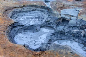 A geothermal mud pool containing thick grey mud, with several connected circular depressions. The surrounding ground is cracked and coloured brown, orange and grey from mineral deposits, creating a rugged volcanic landscape.