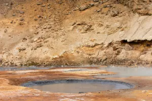 A shallow geothermal pool with mineral-stained ground and rocky earth banks in a barren landscape, showing layers of orange, brown and cream-coloured sediment around the water.